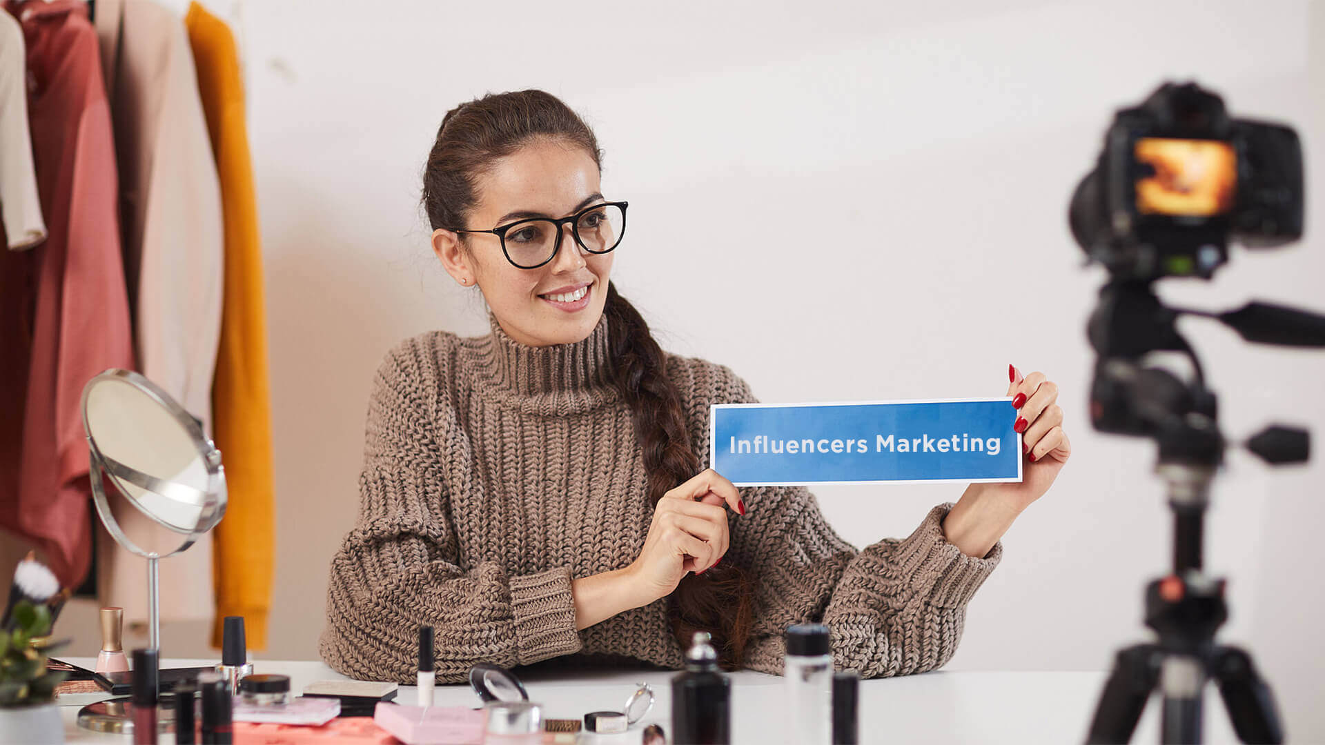 Female influencer in a cozy sweater and glasses holding a sign that says 'Influencers Marketing,' sitting in front of a camera with makeup products, emphasizing local content creation, creative agency involvement, creative advertising, social media viral video production, and social media post strategy.