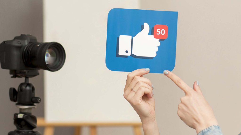 A person holds up a sign with a blue background showing a white thumbs-up icon with a notification bubble displaying the number 50, symbolizing social media engagement. In the background, a camera on a tripod highlights the focus on video marketing strategy for attracting and converting customers.