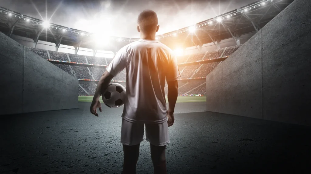 A soccer player stands in a tunnel holding a ball, facing a brightly lit stadium, symbolizing Saudi Arabia's hosting of the 2034 World Cup.